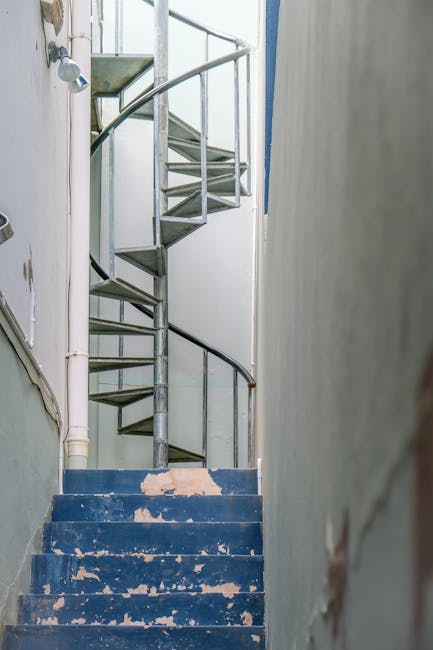 A narrow, spiral metal staircase with open risers and a black handrail ascending in a small, enclosed space. The stairs are located next to a white wall that shows signs of peeling paint, and the step