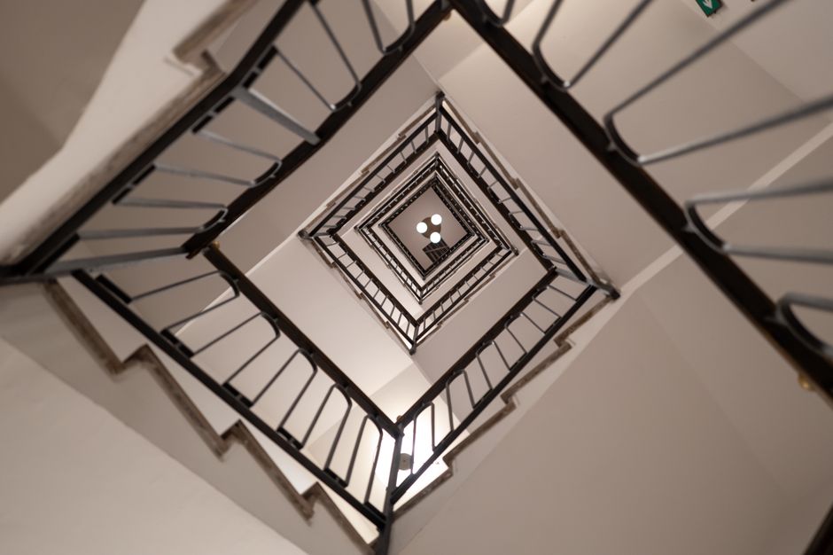 A narrow, spiral metal staircase with open risers and a black handrail ascending in a small, enclosed space. The stairs are located next to a white wall that shows signs of peeling paint, and the step